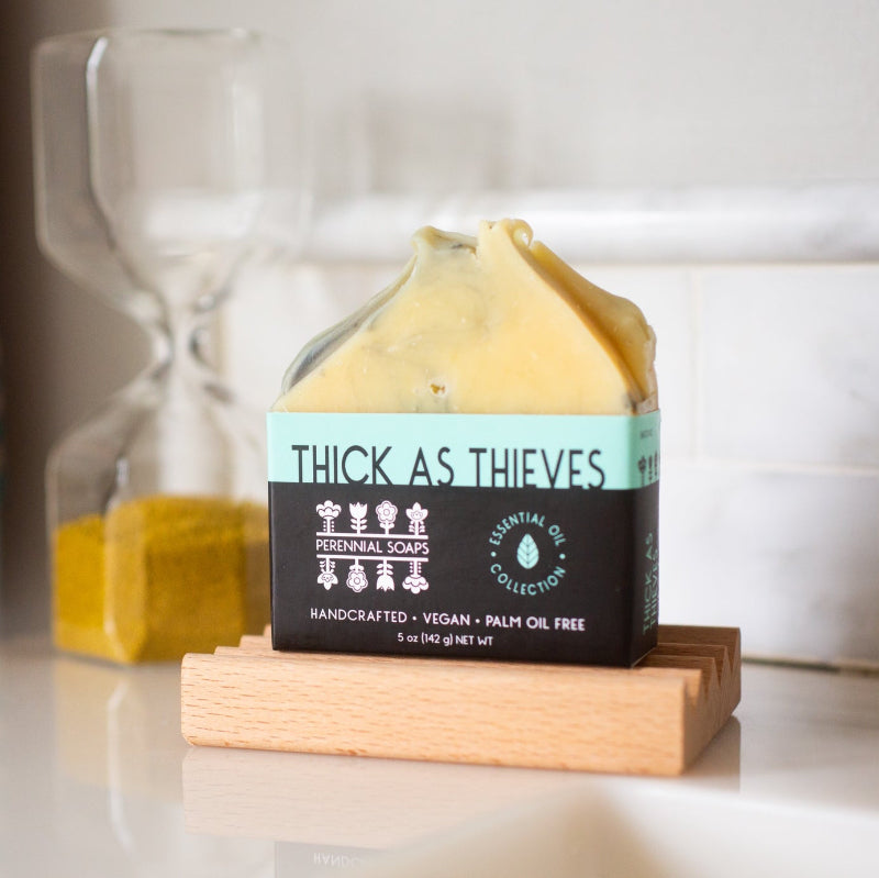 A bar of soap with the label thick as thieves on a wooden soap stand on a white background. 