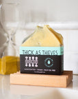 A bar of soap with the label thick as thieves on a wooden soap stand on a white background. 