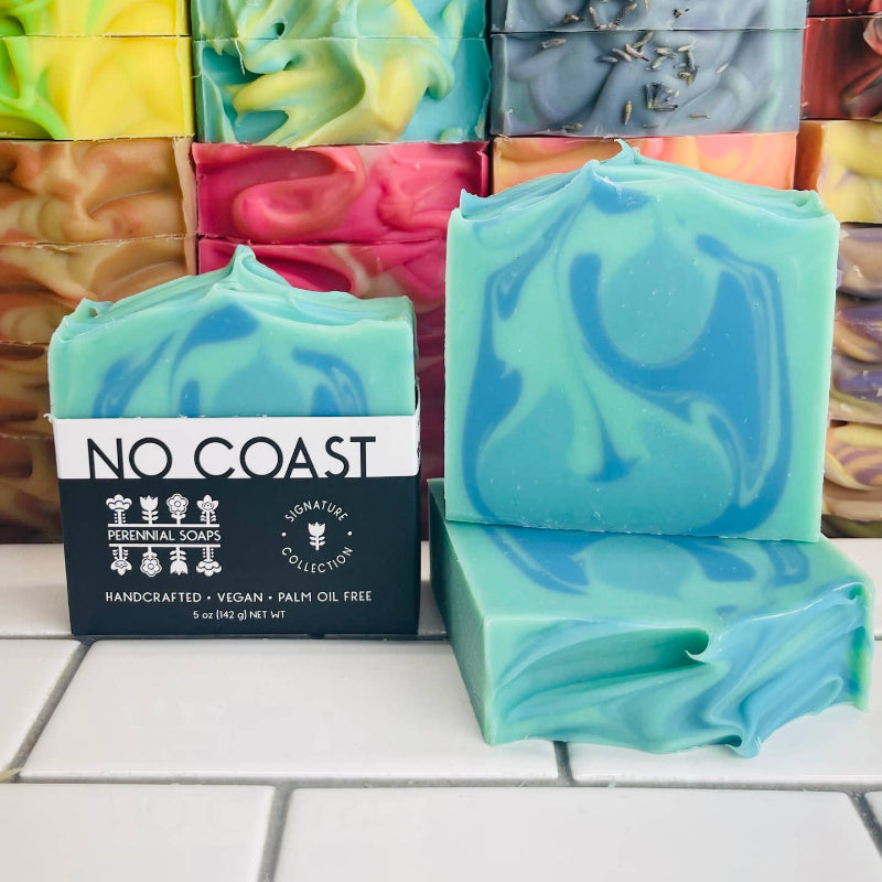 Perennial Soaps No Coast bar sits in-front of a stack of colorful bar soaps. The No Coast bar is light blue with dark blue and sage swirls. 
