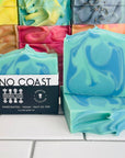 Perennial Soaps No Coast bar sits in-front of a stack of colorful bar soaps. The No Coast bar is light blue with dark blue and sage swirls. 