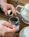 Hands holding a Planet B reusable tea bag folded open and scooping loose leaf tea in. 