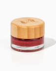 A small glass jar of Zerra & Co. Cherry Flavored Plant Based Jelly with a light wooden lid rests on a white surface. The jelly is red with a shimmer. 