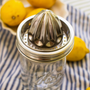 A stainless steel manual citrus juicer with a conical reamer attachment, set on top of a clear glass mason jar. The jar is placed on a blue and white striped cloth with lemons visible in the background.