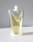 A clear stand-up pouch with a spout cap, filled with light yellow baby wash on a white surface against a light gray background.