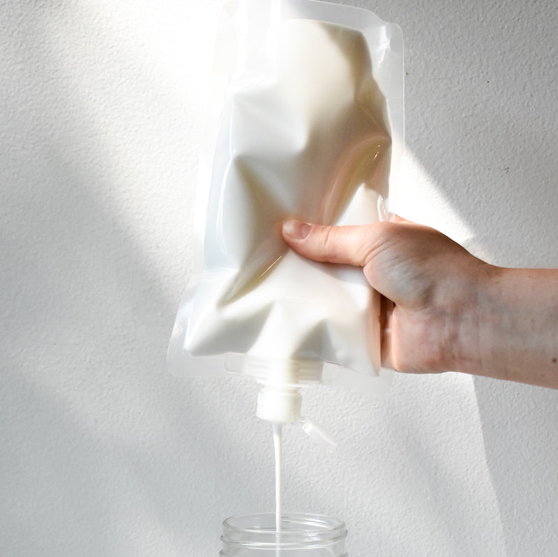 A hand squeezes a white pouch of A Simple Planet Deep Conditioner, pouring the white opaque liquid  into a clear glass jar against a white background.
