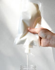 A hand squeezes a white pouch of A Simple Planet Deep Conditioner, pouring the white opaque liquid  into a clear glass jar against a white background.