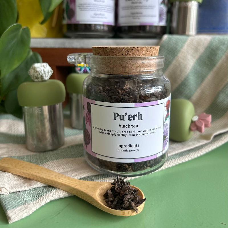 A refillable jar of Pu'erh tea. The jar has a cork lid and an attachable bamboo spoon. the spoon is in front full of tea. 