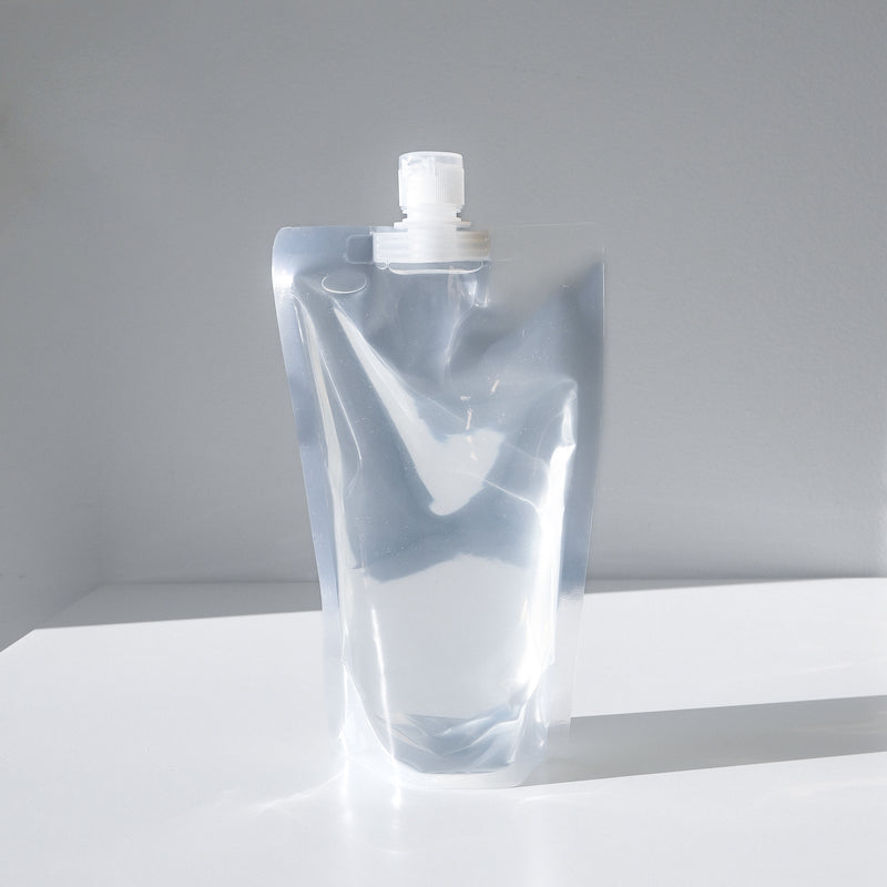 A clear, stand-up plastic pouch with a white screw cap filled with a refillable eyeglasses cleaner sits on a white surface against a light gray background.