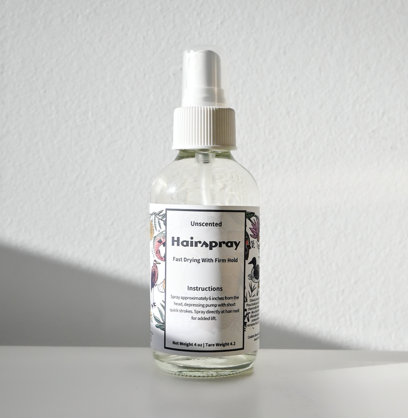 A clear Carina Organics Hairspray bottle with a white label and cap sits on a white surface, casting a shadow. The label displays usage instructions and delicate floral illustrations along the sides.