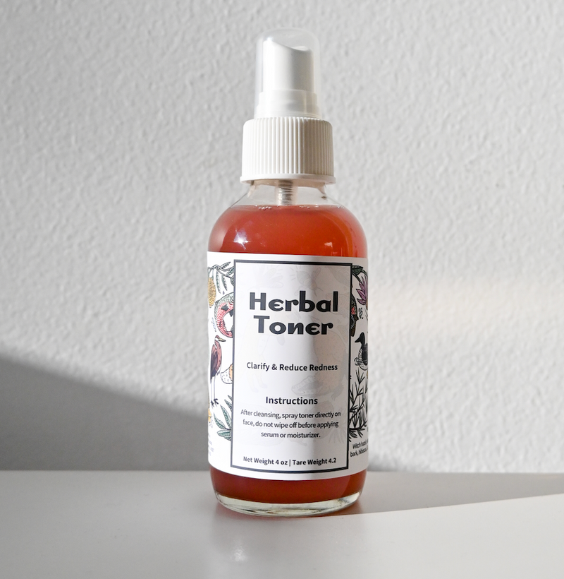 A clear spray bottle labeled Herbal Toner from Green Life Trading Co., filled with a gentle reddish toner, sits on a white surface against a light gray background. The label displays usage instructions and floral illustrations.