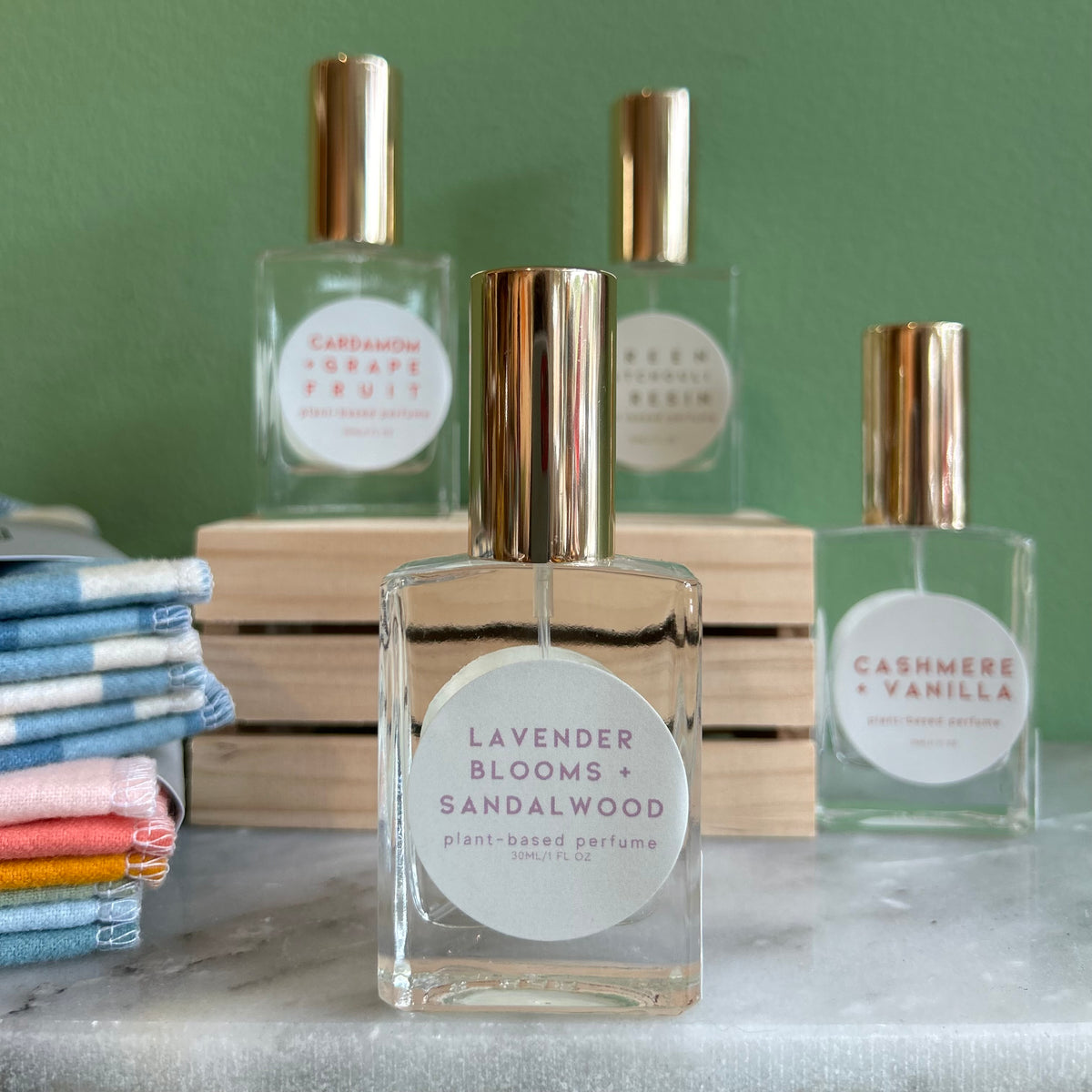 Four glass bottles of refillable vegan perfume with a metallic gold tops on a marble surface and a green back ground.  In front and in focus is a bottle with a circular white label reading "Lavender Blooms + Sandalwood".