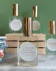 Four glass bottles of refillable vegan perfume with a metallic gold tops on a marble surface and a green back ground.  In front and in focus is a bottle with a circular white label reading "Lavender Blooms + Sandalwood".