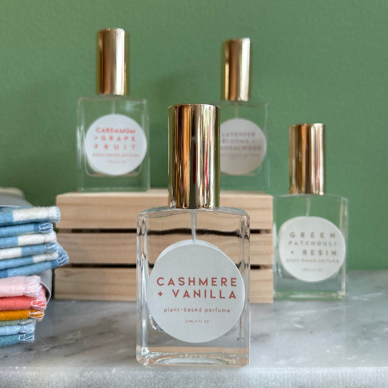 Four glass bottles of refillable vegan perfume with a metallic gold tops on a marble surface and a green back ground.  In front and in focus is a bottle with a circular white label reading "Cashmere + Vanilla".