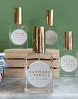 Four glass bottles of refillable vegan perfume with a metallic gold tops on a marble surface and a green back ground.  In front and in focus is a bottle with a circular white label reading "Cashmere + Vanilla".