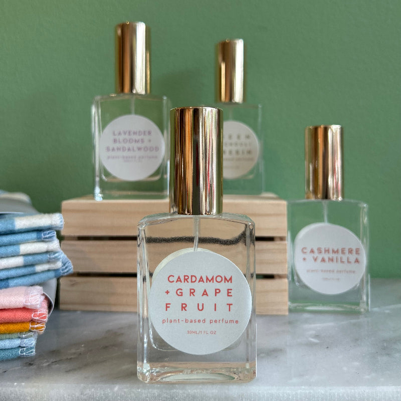 Four glass bottles of refillable vegan perfume with a metallic gold tops on a marble surface and a green back ground.  In front and in focus is a bottle with a circular white label reading "Cardamom + Grapefruit".