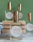 Four glass bottles of refillable vegan perfume with a metallic gold tops on a marble surface and a green back ground.  In front and in focus is a bottle with a circular white label reading "Cardamom + Grapefruit".