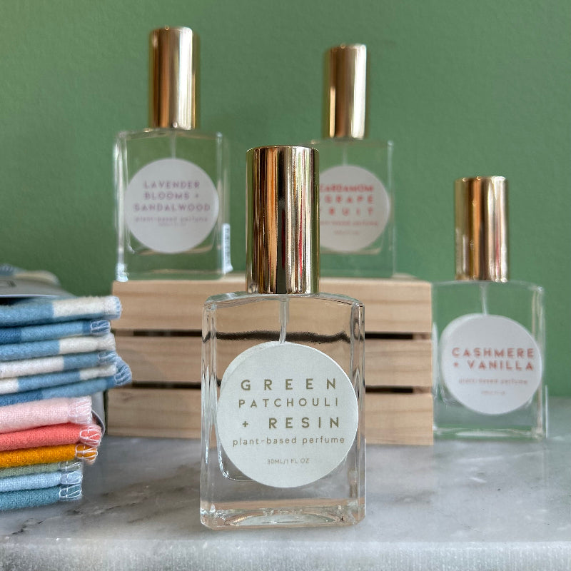 Four glass bottles of refillable vegan perfume with a metallic gold tops on a marble surface and a green back ground.  In front and in focus is a bottle with a circular white label reading "Green Patchouli + Resin".
