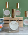 Four glass bottles of refillable vegan perfume with a metallic gold tops on a marble surface and a green back ground.  In front and in focus is a bottle with a circular white label reading "Green Patchouli + Resin".