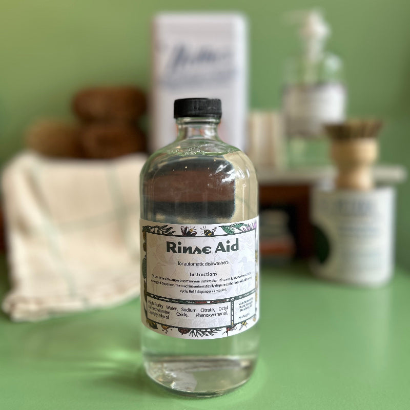 A 16 oz glass bottle of rinse aid that is refillable and reusable. It sits on a green surface with other eco friendly cleaning supplies out of focus behind it.
