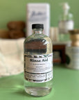 A 16 oz glass bottle of rinse aid that is refillable and reusable. It sits on a green surface with other eco friendly cleaning supplies out of focus behind it.