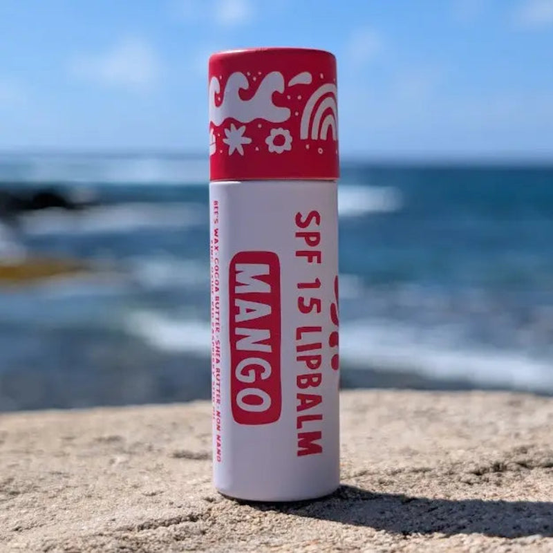 Mango flavored SPF 15 lip balm with red and white paper sustainable packaging upright on a rock on a beach.