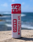 Mango flavored SPF 15 lip balm with red and white paper sustainable packaging upright on a rock on a beach.