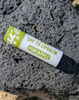 Pineapple flavored SPF 15 lip balm with green and white paper sustainable packaging lying on a rock on a beach.