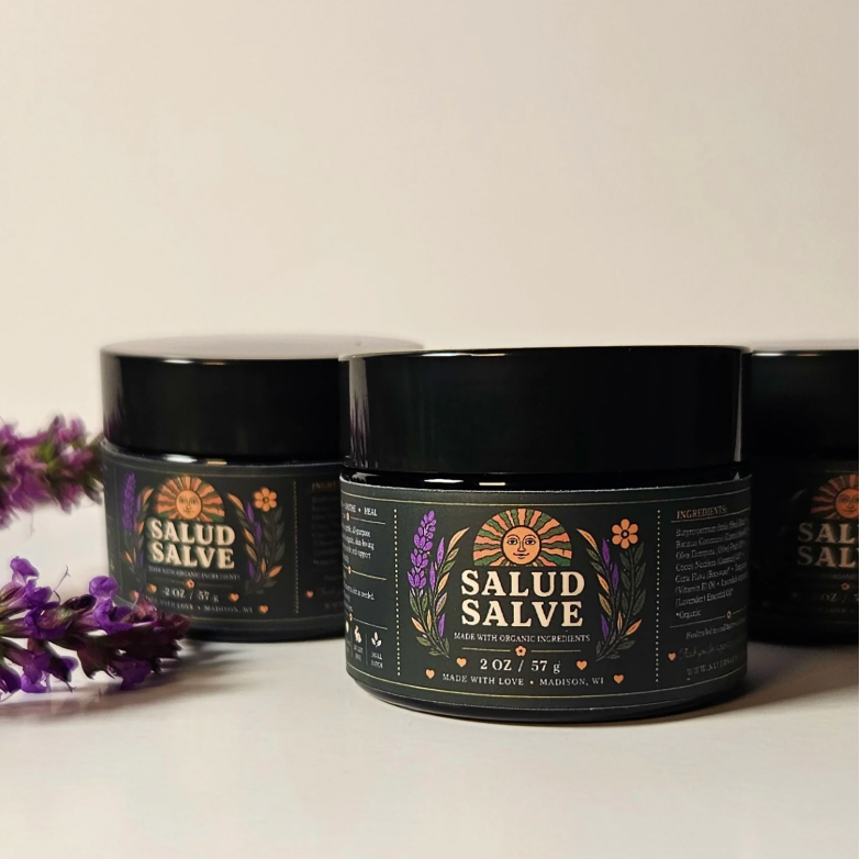 Three black jars of Salud Salve arranged on a light surface with purple lavender sprigs in the foreground. The front jar is in focus, with the other jars softly blurred in the background.