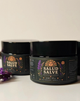 Three black jars of Salud Salve arranged on a light surface with purple lavender sprigs in the foreground. The front jar is in focus, with the other jars softly blurred in the background.