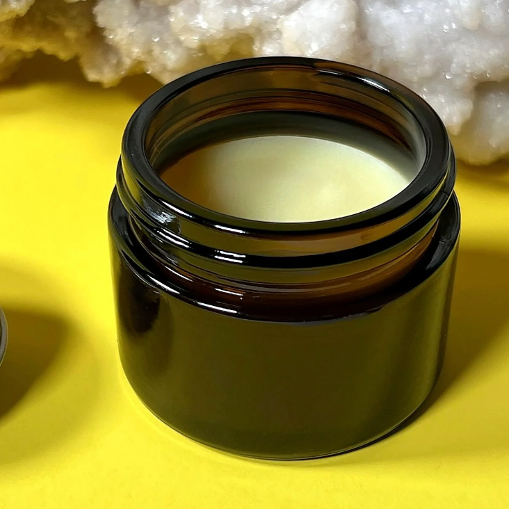 An open dark amber glass jar containing Salud Salve set against a bright yellow background with a white crystal cluster in the upper background.