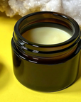 An open dark amber glass jar containing Salud Salve set against a bright yellow background with a white crystal cluster in the upper background.