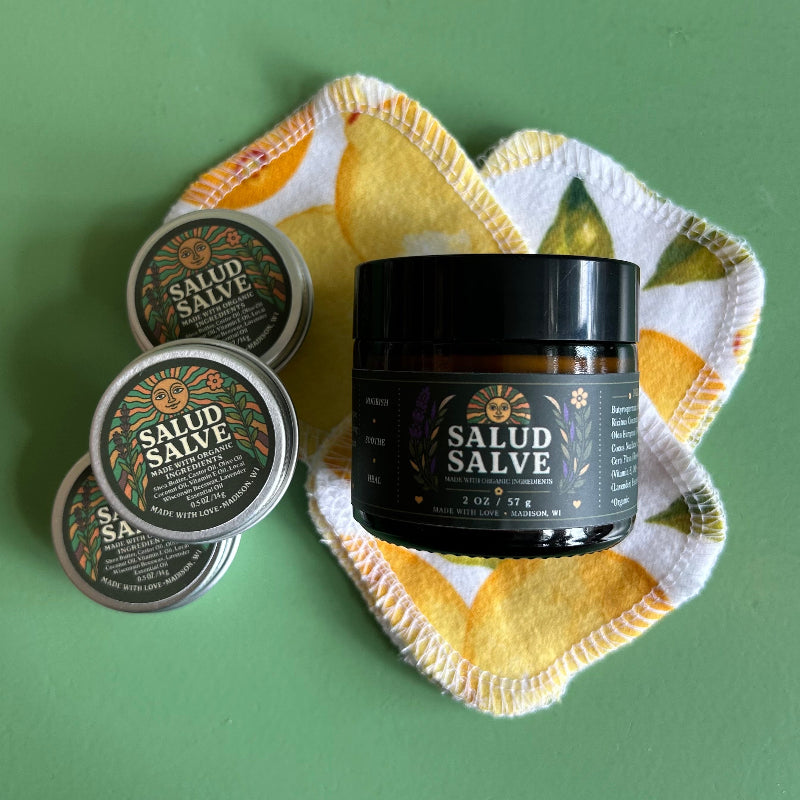 Three tins and a 2 oz amber glass jar of salad salve on top of a green surface with reusable cotton pads.