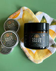 Three tins and a 2 oz amber glass jar of salad salve on top of a green surface with reusable cotton pads.