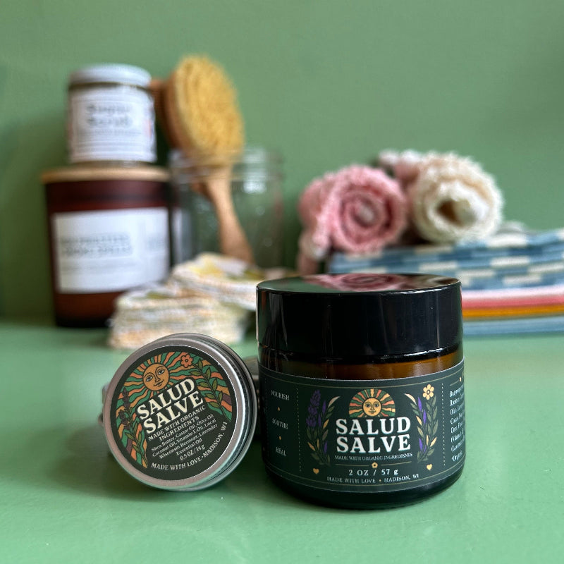 A jar and tin of salud salve with a green background with other eco-friendly goods out of focus behind them. 