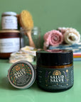 A jar and tin of salud salve with a green background with other eco-friendly goods out of focus behind them. 