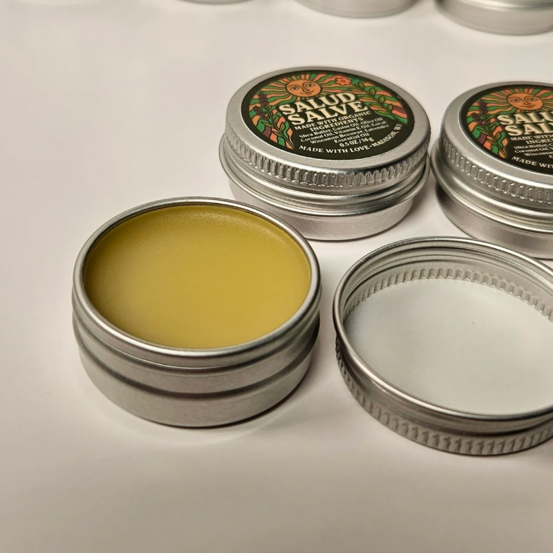 Open silver metal tins of Salud Salve showing the pale yellow salve inside, with one tin closed showing the round label with a sun and lavender, on a white background.