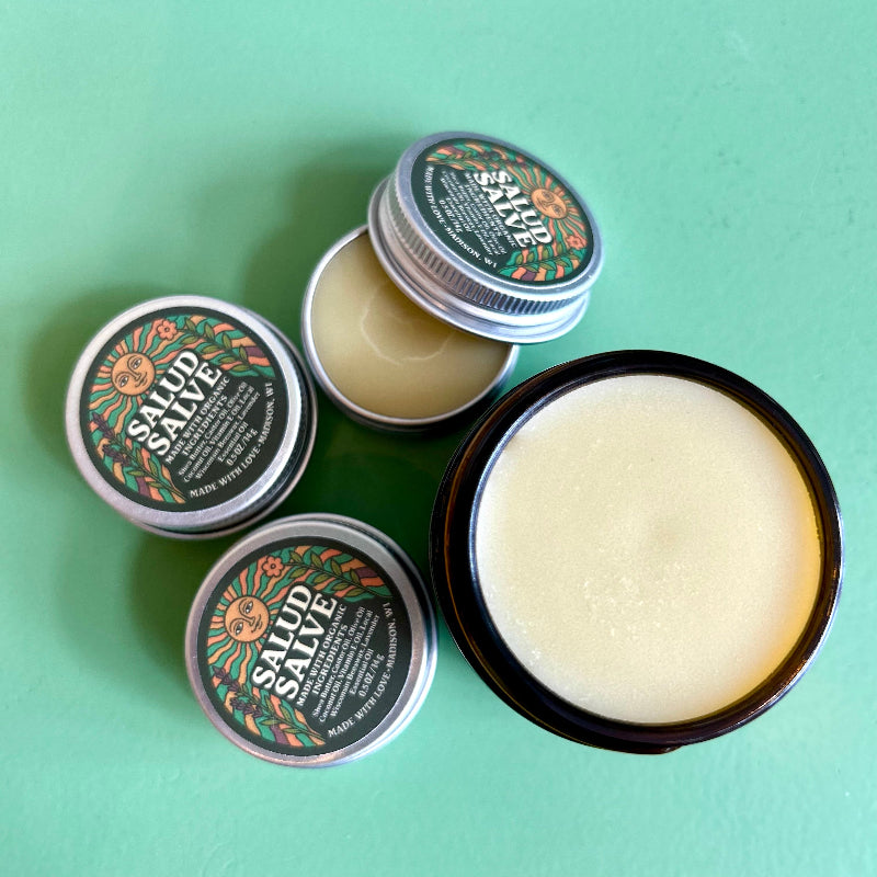 An open jar  and tin of salud salve showing the cream colored nourishing salve inside. 