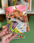 A hand holding a card featuring a high quality print of an oil painting of a fox with its jaw open in a yell. 