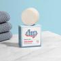 A Dip Gentle Face Wash Bar rests on its blue and white box labeled Gentle Face Wash Bar by Dip atop a marble surface, with folded gray towels in the background—ideal for prepping your face before applying minimal makeup.