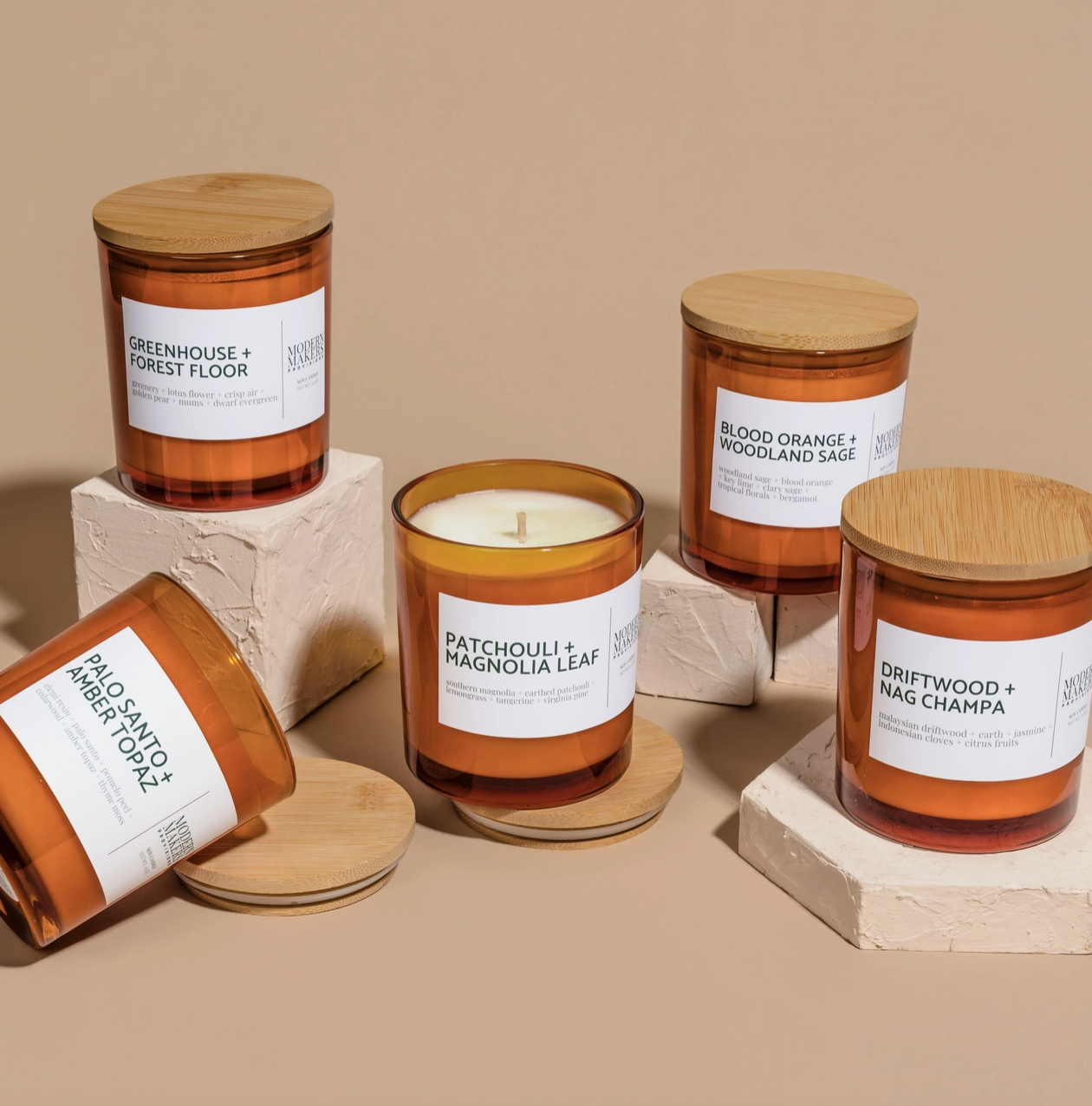 Five Modern Maker Provisions Soy Candles by Modern Makers, each with a wooden lid and unique scent label, are displayed on beige plinths. Hand-poured soy wax promises lasting fragrance; the center candle’s lid is off to reveal the wax inside.