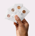 A hand holds four translucent envelopes labeled with circular stickers showing shade numbers 3, 7, 9, and 11. Each envelope contains a sample of Axiology Mushroom Foundation. The background is a plain light color.