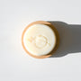 The Cosh Moisturizing Shaving Puck, a round white soap with an embossed minimalist plant design, rests on a wooden dish—ideal as a shave shower tool and casting a soft shadow on a white surface.