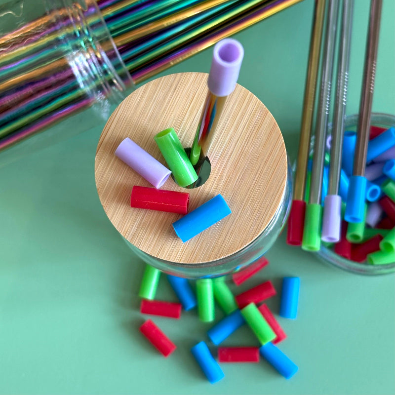 A Bamboo straw lid with a metal straw that has a purple silicone straw tip on it. The lid is surrounded by other silicone reusable straw tips on a green table. 