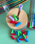 A Bamboo straw lid with a metal straw that has a purple silicone straw tip on it. The lid is surrounded by other silicone reusable straw tips on a green table. 