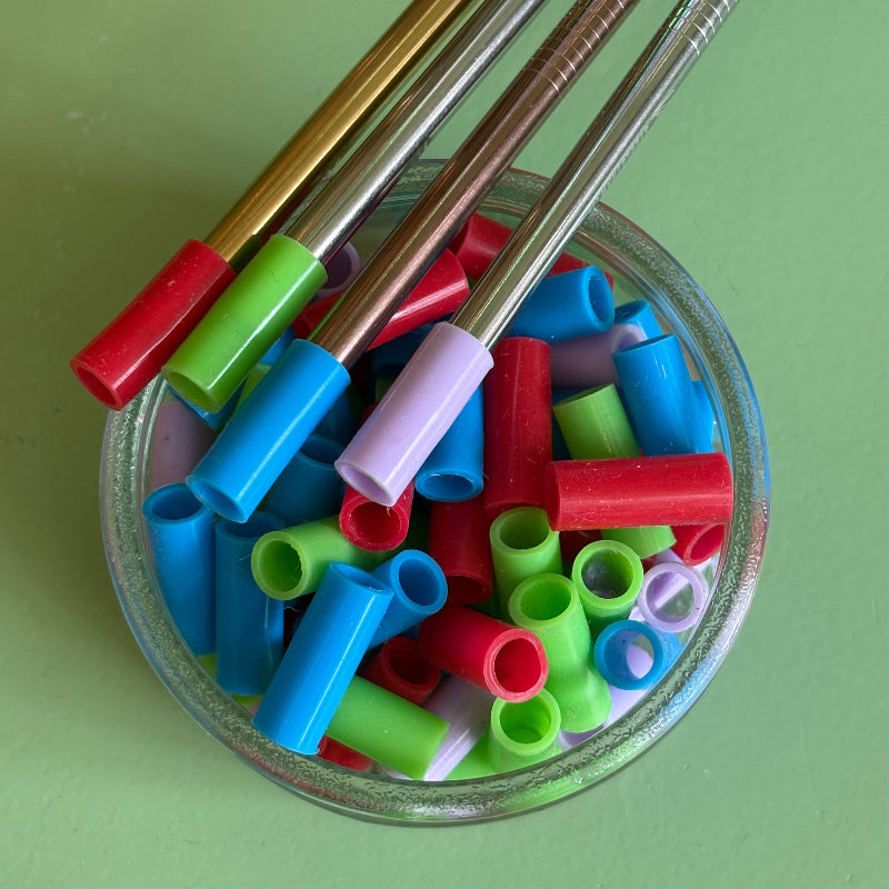 A reused small glass jar full of multicolored reusable silicone straw tips.  Lying on the lip of jar are multicolored metal straws with silicone straw tips on them. 