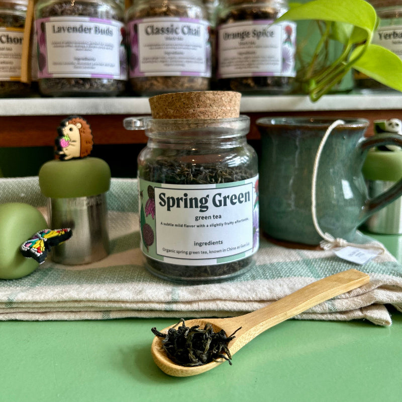 A glass jar of Spring Green tea with a cork lid sits on a green cloth, with a wooden spoon holding loose tea leaves in front. Other tea jars and a ceramic mug are blurred in the background.