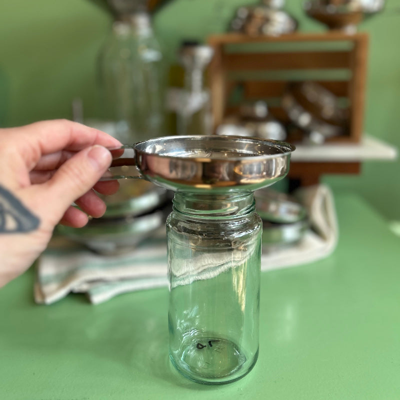 A hand holds a medium sized stainless steel funnel over a small glass jar on a green table top. 