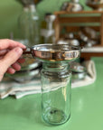 A hand holds a medium sized stainless steel funnel over a small glass jar on a green table top. 