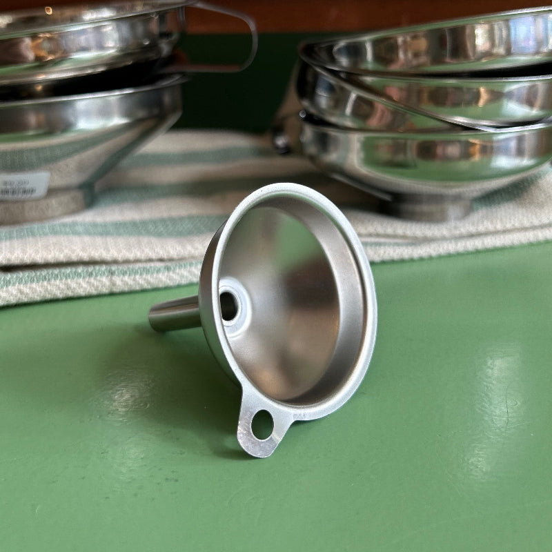 A small stainless steel funnel on a green tabletop with other larger stainless steel funnels in the background. 