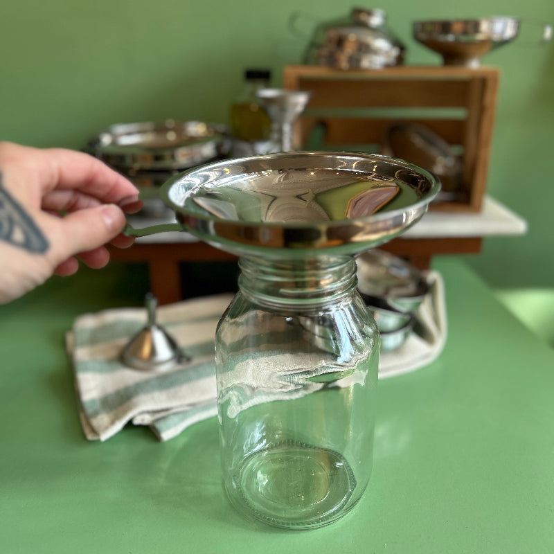 A hand holds a large stainless steel funnel over a large jar on a green table. 